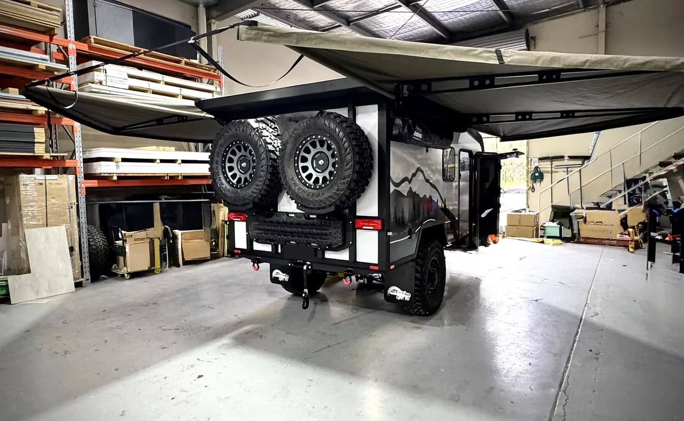 An off-road camper trailer in a workshop, awning out and twin spare wheels on the back.